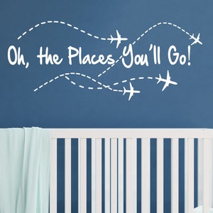 Puede incluir: Pegatina de pared de vinilo blanco con el texto "Oh, the places you'll go!" y una serie de siluetas de aviones con líneas punteadas que representan rutas de vuelo. La pegatina se muestra en una pared azul sobre una cuna blanca.
