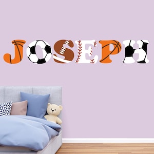 Może przedstawiać: Naklejka ścienna z napisem "JOSEPH" z literami zaprojektowanymi jako piłki sportowe. Litery obejmują koszykówkę, piłkę nożną, futbol amerykański, baseball, koszykówkę i piłkę nożną. Naklejka znajduje się nad łóżkiem.