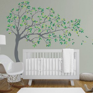 May include: A white crib with a white blanket and a gray tree wall decal with green leaves. The tree branches extend to the right and have leaves that appear to be blowing in the wind.