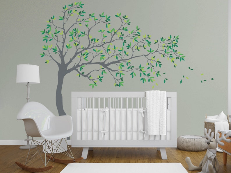 May include: A white crib with a white blanket and a gray tree wall decal with green leaves. The tree branches extend to the right and have leaves that appear to be blowing in the wind.