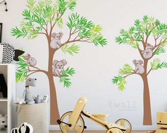 May include: Wall decal featuring two brown trees with green leaves and five gray koalas. The koalas are sitting on the branches of the trees. The text "@wall handmade" is at the bottom of the image.