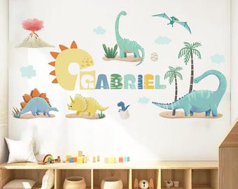 Peut inclure: Un ensemble d'autocollants muraux avec des illustrations de dinosaures et le nom "GABRIEL". Le design comprend divers dinosaures, des palmiers, des nuages et un volcan, créant un thème fantaisiste pour une chambre d'enfant.