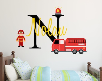 May include: A red fire truck with a black ladder and a yellow siren on top of the letter N. The letter N is black and the name Nolan is written in yellow cursive. A cartoon firefighter in a red uniform stands to the left of the letter N.