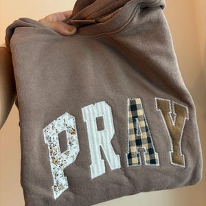 以下が含まれることがあります： 前面に「PRAY」の文字がアップリケされた茶色のパーカー。文字は、花柄、ストライプ、チェック柄、スエードのような質感など、さまざまなパターンになっています。パーカーは折りたたまれ、手で持たれています。