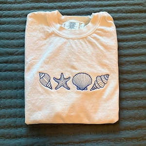 Może przedstawiać: Biała koszulka z niebieskimi haftowanymi muszelkami i gwiazdą morską. Koszulka ma metkę "Comfort Colors".