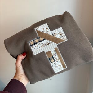 以下が含まれることがあります： パッチワークの十字架のデザインが施された茶色のスウェットシャツ。十字架は、花柄、チェック柄、無地の茶色の部分など、さまざまな生地で作られています。スウェットシャツは折りたたまれています。