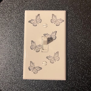 Könnte beinhalten: Eine weiße Lichtschalterabdeckung, verziert mit sechs Schmetterlingsillustrationen. Der Schalter befindet sich in der Position "ON". Die Schmetterlinge sind in einem silbernen Glitzerdesign gehalten. Die Abdeckung ist rechteckig und hat zwei Schrauben.