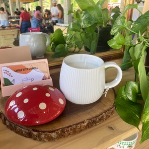 May include: White ceramic mug candle with a ribbed design and a handle. The candle is on a wooden slice with a red and white polka dot mushroom.