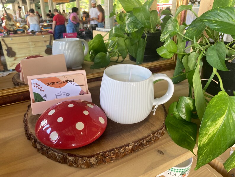 May include: White ceramic mug candle with a ribbed design and a handle. The candle is on a wooden slice with a red and white polka dot mushroom.