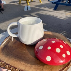 May include: A white ceramic mug with a ribbed design and a red and white polka dot ceramic mushroom lid. The mug and lid are sitting on a wooden slice.