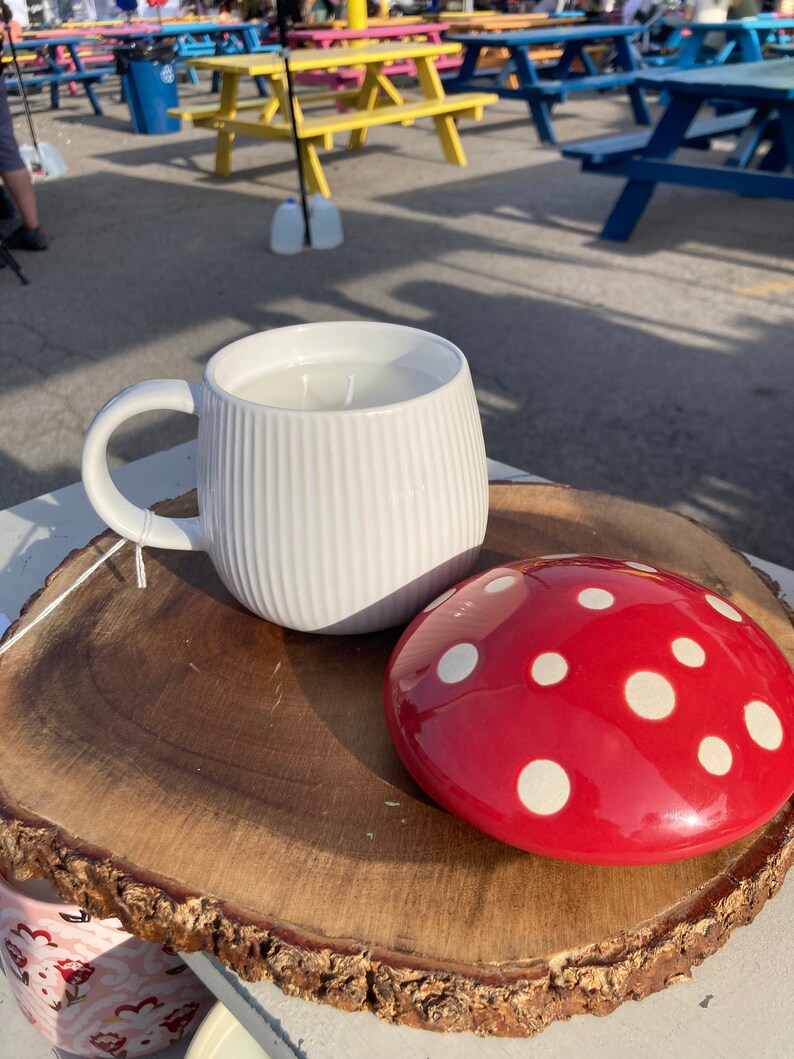 May include: A white ceramic mug with a ribbed design and a red and white polka dot ceramic mushroom lid. The mug and lid are sitting on a wooden slice.
