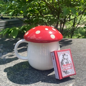 May include: A white ceramic mug with a red and white mushroom lid. The mug has a white and red striped design. The mug is sitting on a gray surface next to a red and white book with the title "Alice's Adventures in Wonderland".