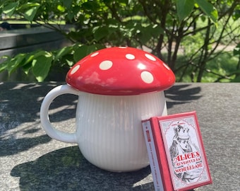 May include: A white ceramic mug with a red and white mushroom lid. The mug has a white and red striped design. The mug is sitting on a gray surface next to a red and white book with the title "Alice's Adventures in Wonderland".