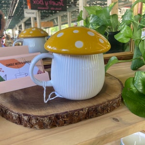 May include: White ceramic mug with a yellow and white mushroom lid. The mug has a ribbed design and a handle. The mug is sitting on a wooden slice.