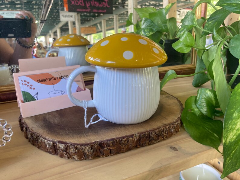 May include: White ceramic mug with a yellow and white mushroom lid. The mug has a ribbed design and a handle. The mug is sitting on a wooden slice.