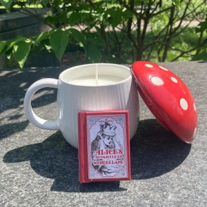 May include: White ceramic mug with a red and white polka dot lid, a white candle inside, and a small red book with the title "Alice's Adventures in Wonderland" on the front.