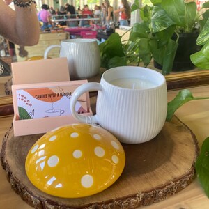May include: A white ceramic candle with a ribbed design and a handle. The candle is on a wooden slice with a yellow and white polka dot mushroom-shaped lid.
