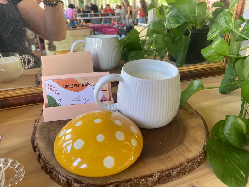 May include: A white ceramic candle with a ribbed design and a handle. The candle is on a wooden slice with a yellow and white polka dot mushroom-shaped lid.