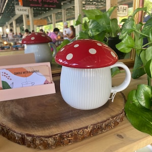 May include: White ceramic mug with a red and white mushroom lid. The mug has a handle and vertical lines on the side. The mug is sitting on a wooden slice.