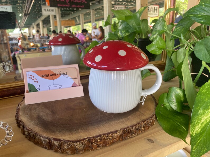 May include: White ceramic mug with a red and white mushroom lid. The mug has a handle and vertical lines on the side. The mug is sitting on a wooden slice.