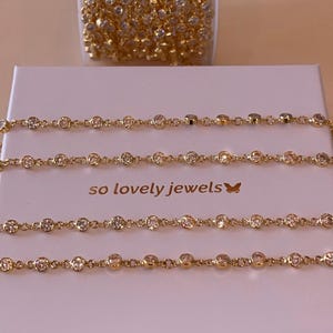 Peut inclure: Une chaîne dorée avec des pierres rondes transparentes serties, disposée sur une boîte carrée blanche. La boîte porte l'inscription "so lovely jewels" accompagnée d'une petite icône de papillon. Une bobine de chaîne similaire est posée au-dessus.