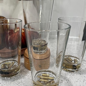 May include: Clear glass tumblers with brass bullet casings embedded in the base. The glasses vary in size and shape, with a shot glass and a small wooden container in the background. The arrangement suggests a unique barware set.