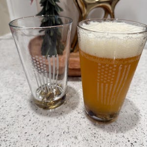 May include: Two clear pint glasses with a gold base, one empty and one filled with a golden liquid and foam. Each glass is etched with a design resembling the American flag. The glasses are on a speckled white surface.