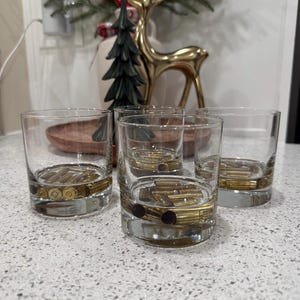 May include: Four clear, short whiskey glasses with a band of brass bullet casings embedded in the base. The glasses are arranged on a speckled countertop, with a golden deer figurine and Christmas decor in the background.