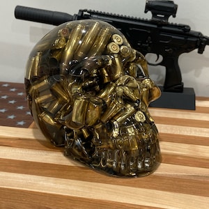 May include: A resin skull filled with brass bullet casings. The skull is positioned in front of a black firearm with a red dot sight. The skull and firearm are on a wooden surface with a striped pattern.
