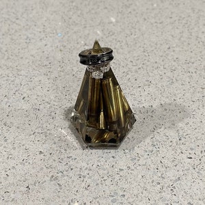 May include: Two black wedding bands with diamond accents are displayed on a clear glass ring holder filled with gold-colored bullet casings.