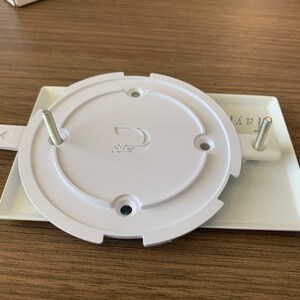 Puede incluir: Placa de pared blanca de pl&aacute;stico con dos tornillos y una tapa redonda blanca con un logotipo que dice "D" y un peque&ntilde;o dise&ntilde;o de puntos. La placa tiene la palabra "AAR" impresa.