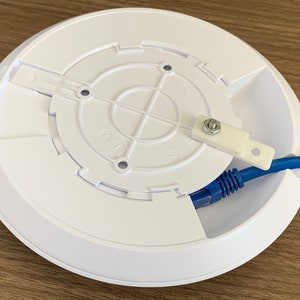 Puede incluir: Soporte de techo blanco para un router inal&aacute;mbrico con un cable Ethernet azul conectado. El soporte tiene una abertura circular con cuatro agujeros para tornillos y una peque&ntilde;a leng&uuml;eta de pl&aacute;stico con un tornillo.