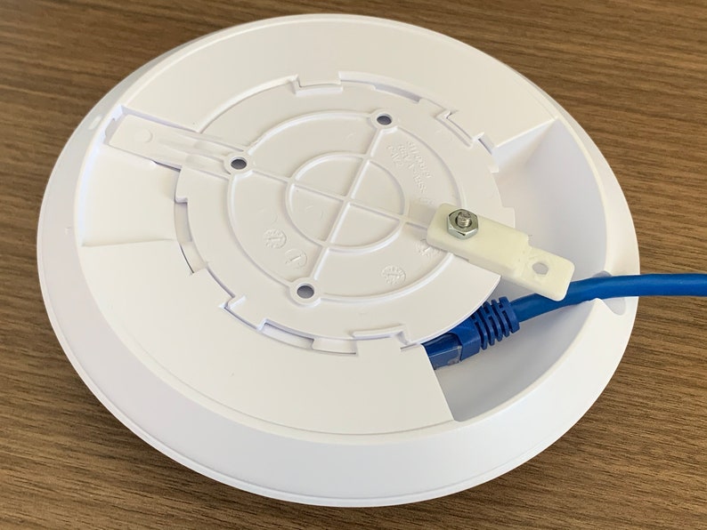 Puede incluir: Soporte de techo blanco para un router inal&aacute;mbrico con un cable Ethernet azul conectado. El soporte tiene una abertura circular con cuatro agujeros para tornillos y una peque&ntilde;a leng&uuml;eta de pl&aacute;stico con un tornillo.