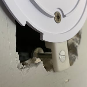 Puede incluir: Primer plano de una caja el&eacute;ctrica de pl&aacute;stico blanco que se est&aacute; instalando en una pared. La caja est&aacute; asegurada con un tornillo y un soporte met&aacute;lico. La pared est&aacute; hecha de yeso y est&aacute; agrietada alrededor del agujero donde se est&aacute; instalando la caja.