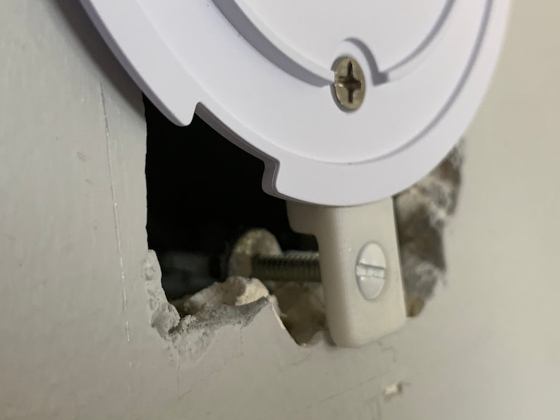 Puede incluir: Primer plano de una caja el&eacute;ctrica de pl&aacute;stico blanco que se est&aacute; instalando en una pared. La caja est&aacute; asegurada con un tornillo y un soporte met&aacute;lico. La pared est&aacute; hecha de yeso y est&aacute; agrietada alrededor del agujero donde se est&aacute; instalando la caja.