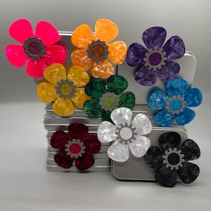 Peut inclure: Assortiment de médiators de guitare en forme de fleurs colorées avec un motif d'engrenage au centre. Les médiators sont disposés en cercle sur une boîte en étain argentée.