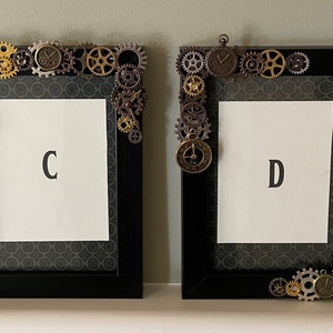 Puede incluir: Dos marcos de fotos negros con un dise&ntilde;o steampunk. Los marcos est&aacute;n decorados con engranajes, pi&ntilde;ones y esferas de reloj. Los marcos est&aacute;n etiquetados con "C" y "D".
