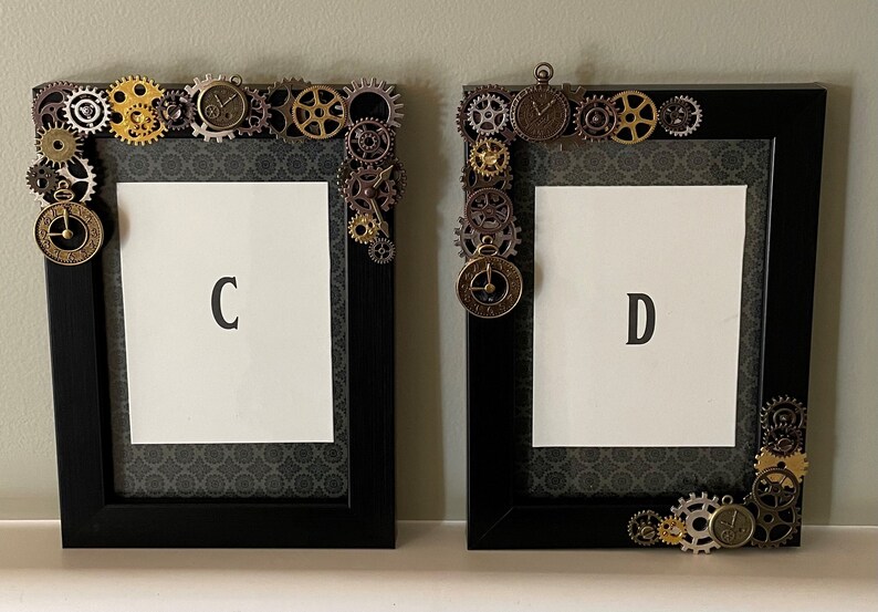 Puede incluir: Dos marcos de fotos negros con un dise&ntilde;o steampunk. Los marcos est&aacute;n decorados con engranajes, pi&ntilde;ones y esferas de reloj. Los marcos est&aacute;n etiquetados con "C" y "D".