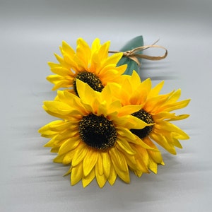 May include: A bouquet of three yellow sunflowers with dark brown centers. The sunflowers are arranged in a bunch and tied together with twine.