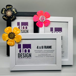 May include: Three white picture frames with black mats, each decorated with a different colorful paper flower. The frames are stacked on top of each other, with the largest frame on the bottom. The smallest frame is on top. The text "4 x 6 FRAME" is printed on the front of the middle frame, along with the company's website and social media handles.