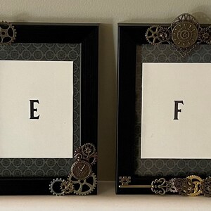 Puede incluir: Dos marcos de fotos negros con adornos met&aacute;licos ornamentados, incluyendo engranajes y una llave. Los marcos est&aacute;n decorados con un fondo de patr&oacute;n gris oscuro. El marco de la izquierda tiene la letra "E" en el centro y el marco de la derecha tiene la letra "F" en el centro.