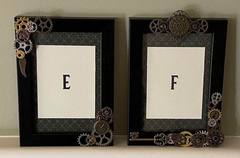 Puede incluir: Dos marcos de fotos negros con adornos met&aacute;licos ornamentados, incluyendo engranajes y una llave. Los marcos est&aacute;n decorados con un fondo de patr&oacute;n gris oscuro. El marco de la izquierda tiene la letra "E" en el centro y el marco de la derecha tiene la letra "F" en el centro.