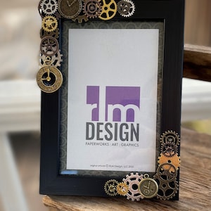 Puede incluir: Marco de imagen negro con un logotipo morado y blanco que dice "rm DESIGN PAPERWORKS : ART : GRAPHICS" y el texto "original artwork &copy; RLM Design, LLC 2020". El marco est&aacute; decorado con engranajes dorados y plateados y esferas de reloj.