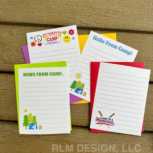 May include: A collection of colorful camp-themed notepads. Each notepad features a different design, including text that reads "Summer Camp News," "News From Camp," "Hello From Camp," and "Camp is a Homerun." The notepads have lined pages.