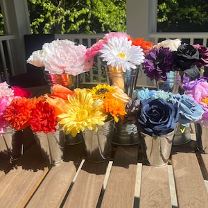 May include: A collection of colorful artificial flowers arranged in silver buckets. The flowers include pink peonies, yellow gerbera daisies, orange roses, purple hydrangeas, and blue roses.