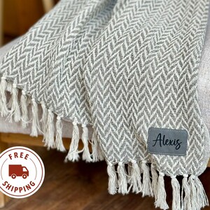 May include: A gray and white herringbone throw blanket with fringe on the edges. The blanket is draped over a chair and has a personalized tag that says "Alexis".