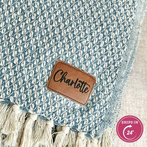 May include: A blue and white patterned throw blanket with fringe. The blanket has a leather tag with the name "Charlotte" on it.