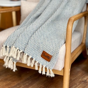 May include: A blue and white woven throw blanket with fringe on the edges. The blanket is draped over a wooden chair and has a brown leather tag with the name "Charlotte" on it.