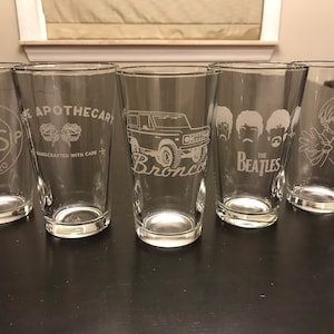May include: Five clear glass pint glasses with various designs etched into the glass. The designs include "The Pint N Sip Est. 2020", "The Apothecary Handcrafted with Care", "Bronco", "The Beatles", and a cartoon character.