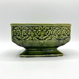 Puede incluir: Una jardinera de cerámica verde con forma rectangular y base de pedestal. La jardinera presenta un diseño ornamental de volutas repetidas alrededor de los lados. El acabado brillante le da un aspecto vintage.